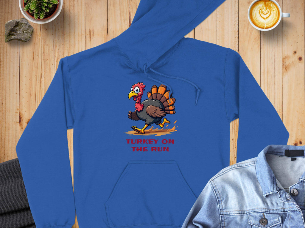 A blue hoodie featuring a cartoon turkey and Turkey On The Run by Garment Graphics sprawls across a wooden table, complemented by a denim jacket, latte, and plant in this cozy scene.