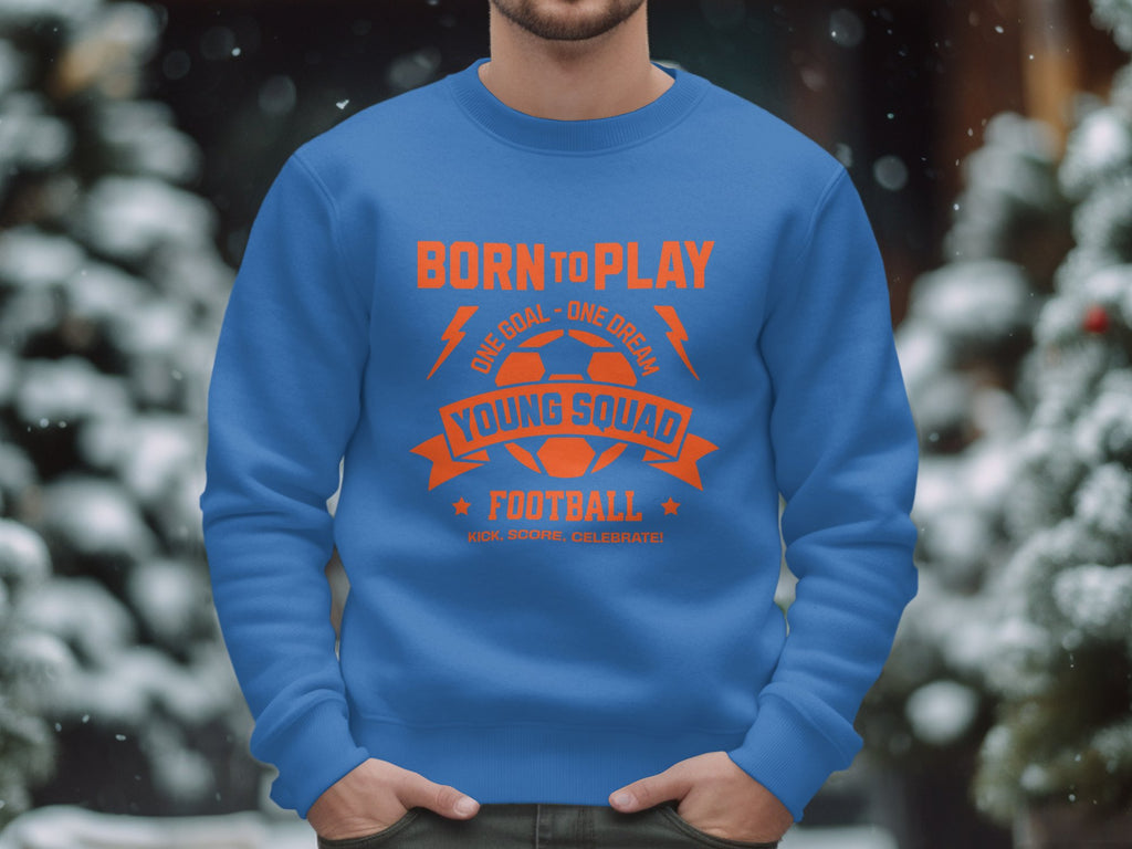 A person wearing a blue Garment Graphics football sweatshirt stands outdoors. This unisex design, featuring the words Born to Play, Young Squad, and Football in bold orange letters, is perfect for young football enthusiasts. Snow-covered trees are visible in the background.