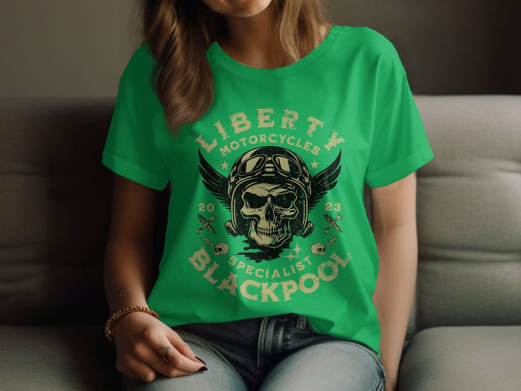 A woman sits wearing a Garment Graphics green T-shirt with a skull, wings, and crossed tools design. It features the text Liberty Motorcycles Specialist Blackpool 2023, embodying the essence of Blackpool-themed shirts.