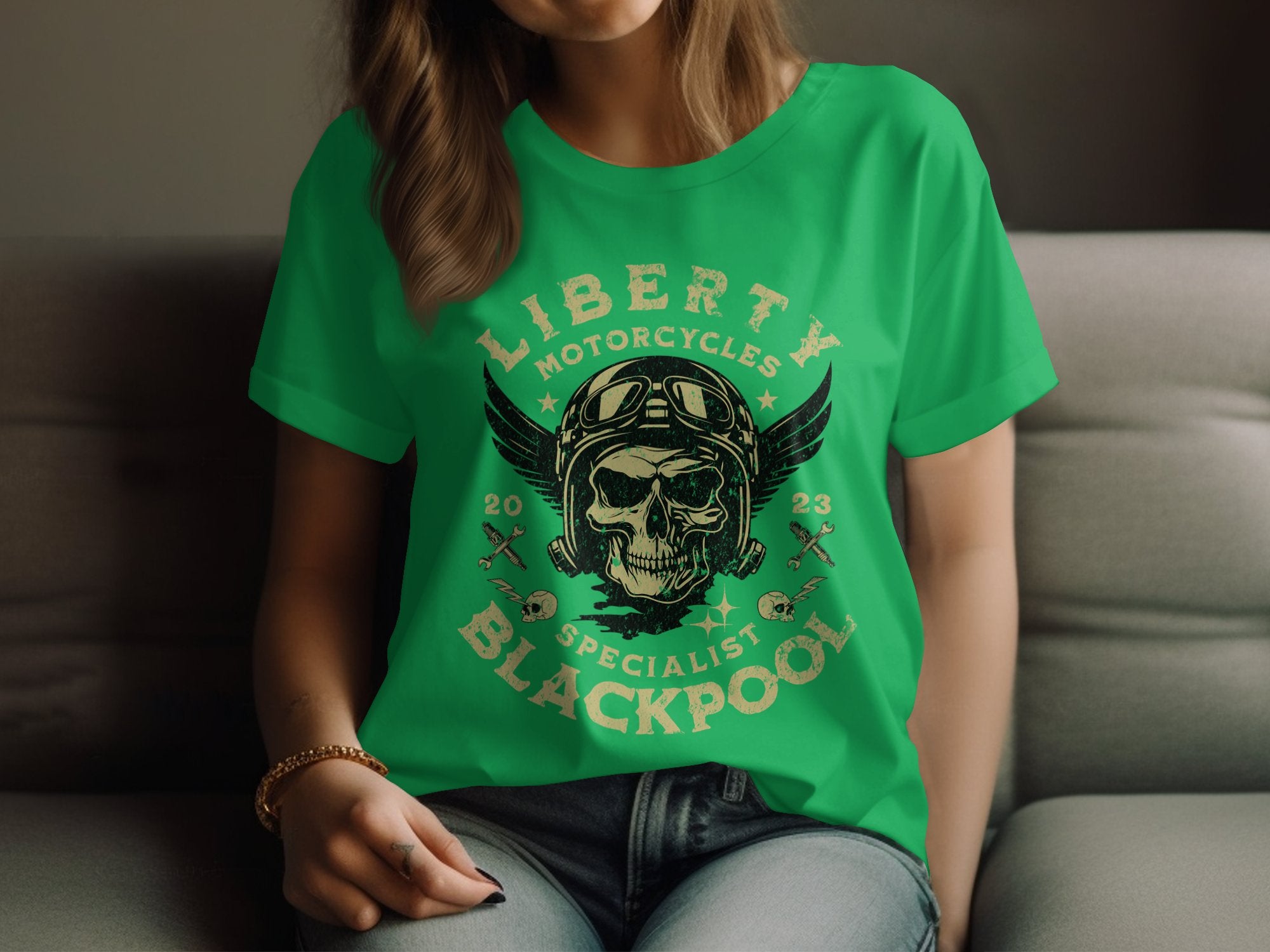 A woman sits wearing a Garment Graphics green T-shirt with a skull, wings, and crossed tools design. It features the text Liberty Motorcycles Specialist Blackpool 2023, embodying the essence of Blackpool-themed shirts.