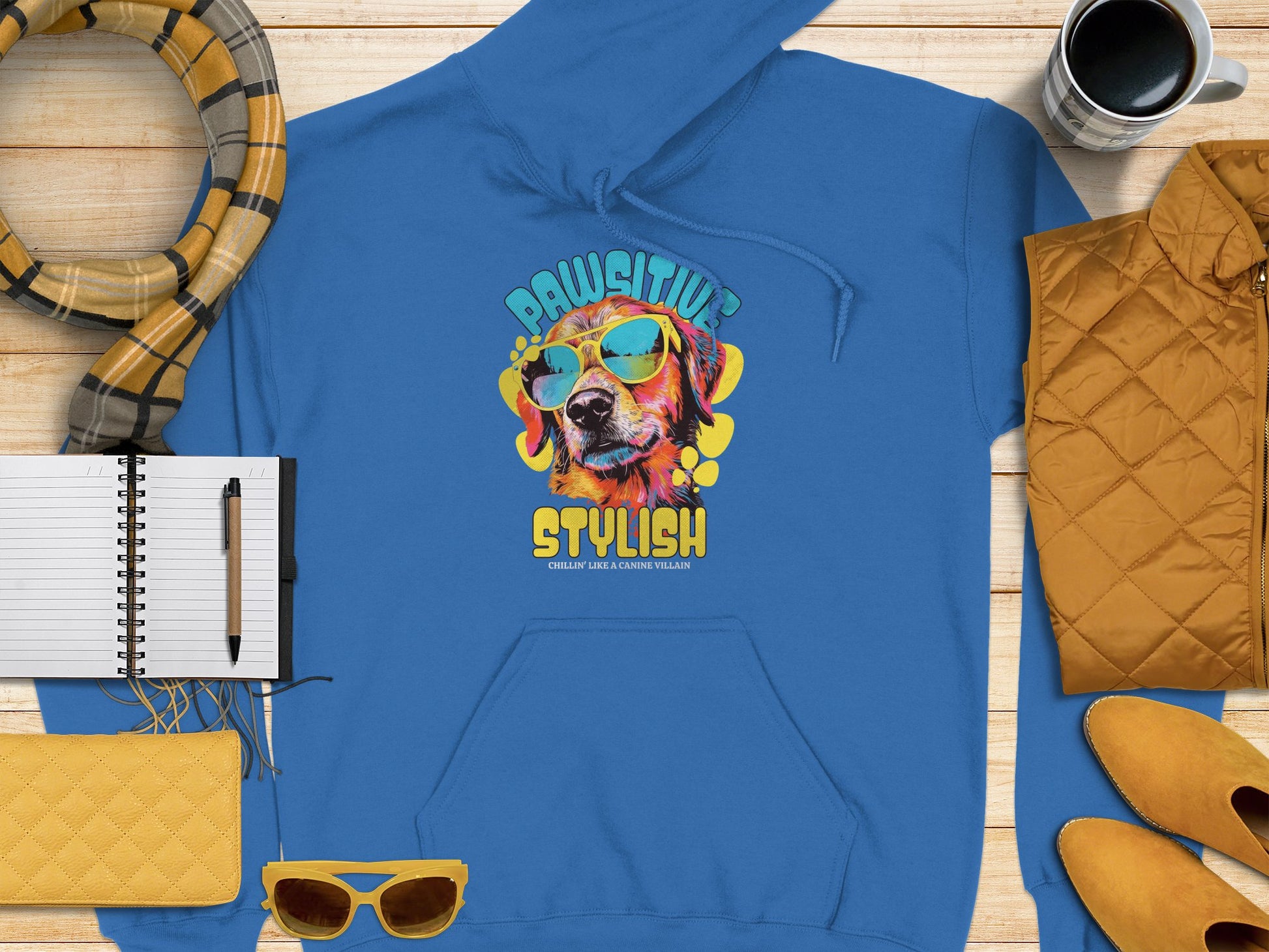 A blue hoodie from Garment Graphics, showcasing a dog in sunglasses with a vibrant yellow and blue design, reading Pawsitively Stylish, is displayed on wood next to a notebook, scarf, quilted yellow items, sunglasses, and coffee.