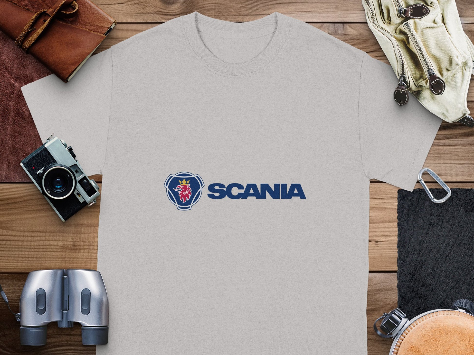 A gray cotton T-shirt from Garment Graphics, featuring a blue emblem with a red griffin head of the Scania logo, is elegantly placed on a wooden surface. Nearby are companion items such as a leather bag, camera, binoculars, compass, and gloves.
