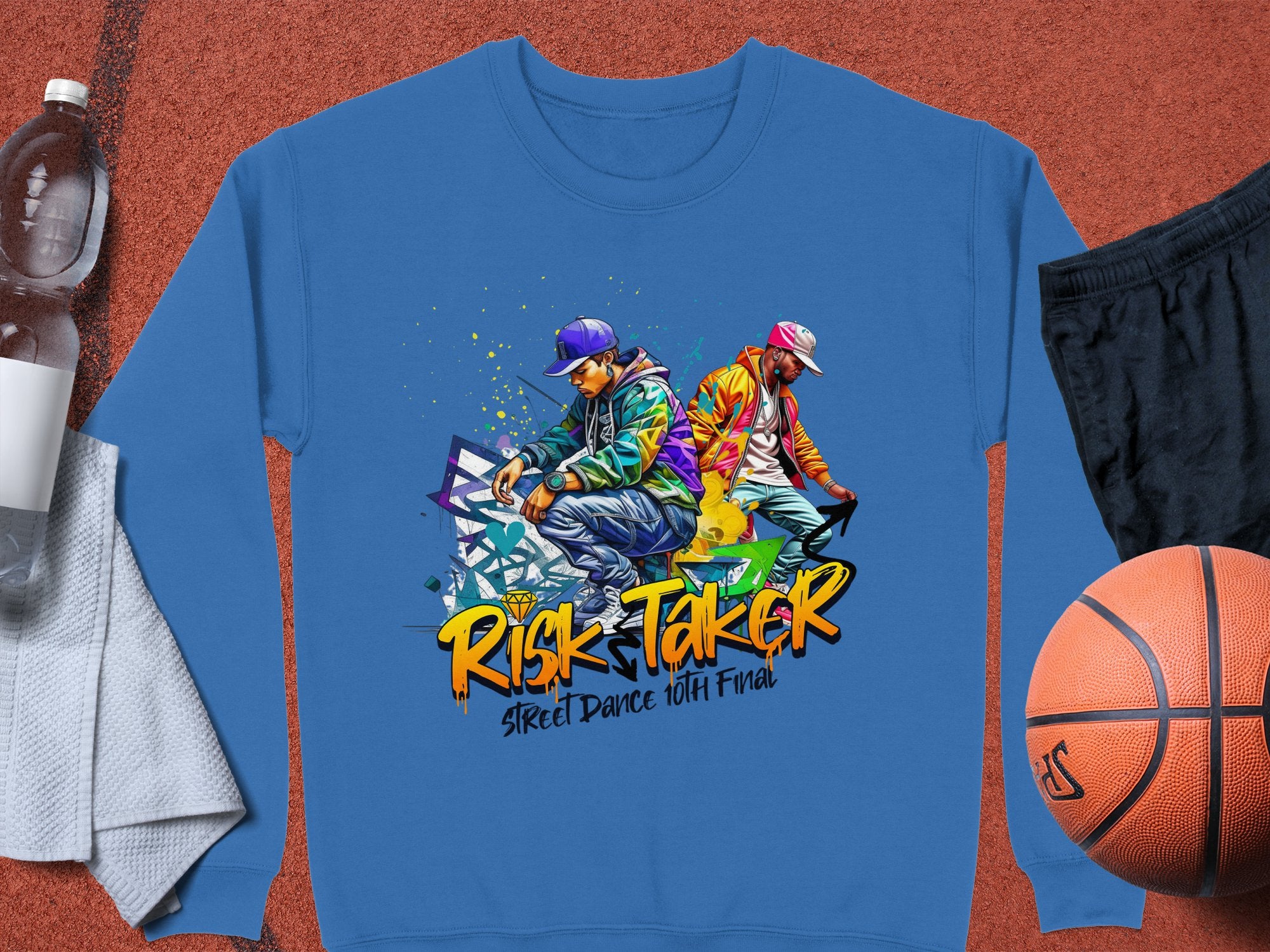 Garment Graphics offers a blue sweatshirt with urban street dance graphics of two vibrant dancers and the text Risk Taker Street Dance 10th Final. It includes a basketball, folded dark shorts, water bottle, and towel. Available in unisex sizing for all street dance enthusiasts.