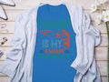 The Garment Graphics blue fishing t-shirt features Fishing is my anger management in green and orange. It displays a fish biting a lure against a wooden surface, paired with a gray cardigan, flowers, and accessories—a must-have for fishing enthusiasts.