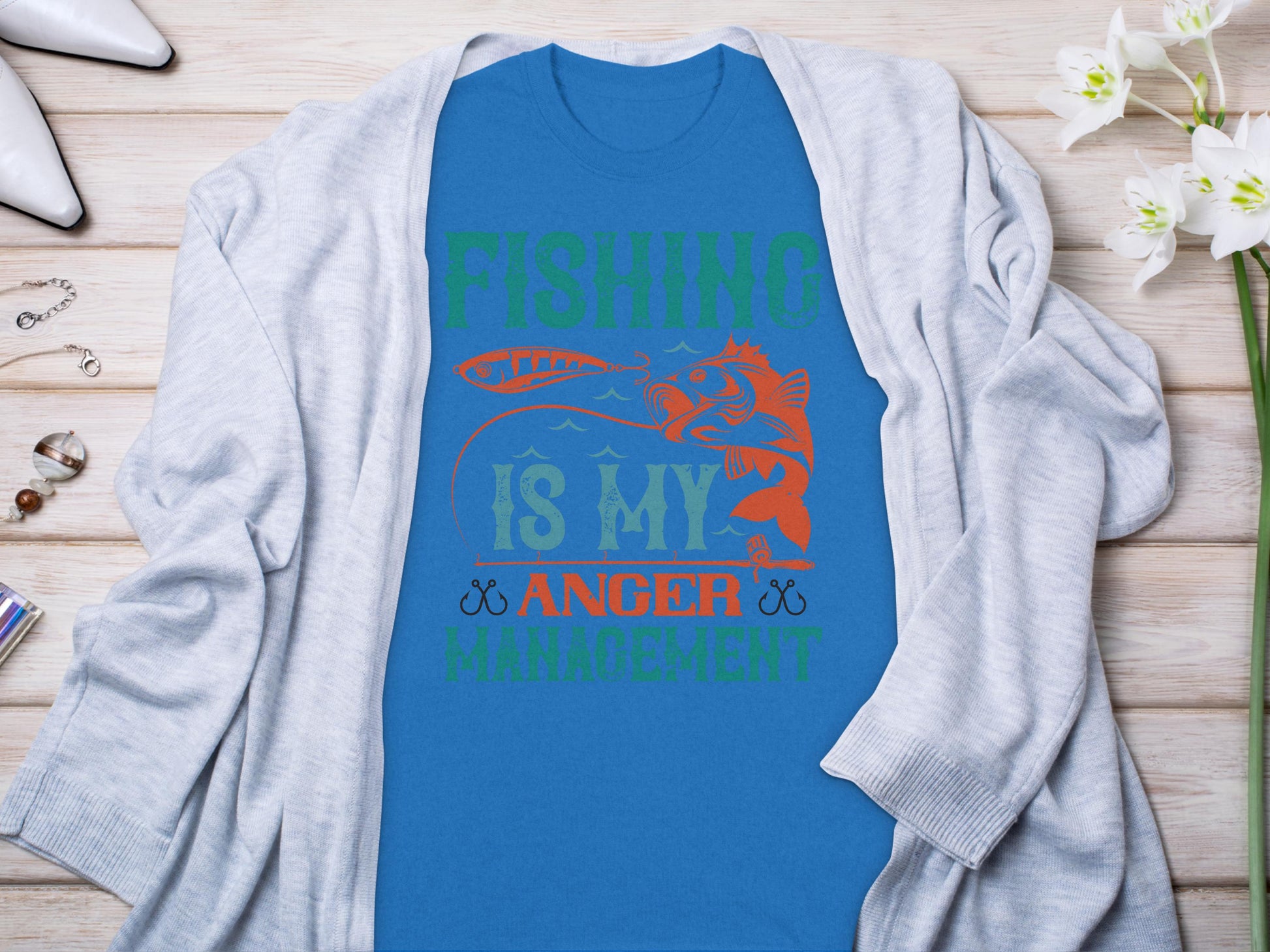 The Garment Graphics blue fishing t-shirt features Fishing is my anger management in green and orange. It displays a fish biting a lure against a wooden surface, paired with a gray cardigan, flowers, and accessories—a must-have for fishing enthusiasts.