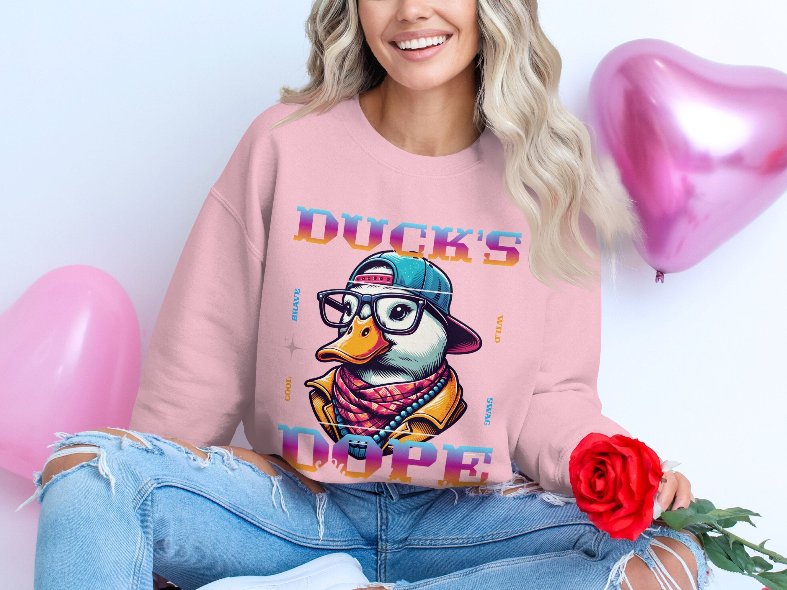 Dressed in a bold pink sweatshirt with a trendy cartoon graphic of Ducks Dope by Garment Graphics, complete with glasses and a cap, they pair it with ripped jeans while holding a red rose amidst pink heart-shaped balloons.