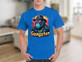A person in a blue Garment Graphics T-Shirt, featuring urban style and a cartoon gorilla with sunglasses, hat, and gold chain. Gangster is boldly printed below. The classic fit pops against the blurry indoor background with light brown flooring.