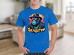 A person in a blue Garment Graphics T-Shirt, featuring urban style and a cartoon gorilla with sunglasses, hat, and gold chain. Gangster is boldly printed below. The classic fit pops against the blurry indoor background with light brown flooring.