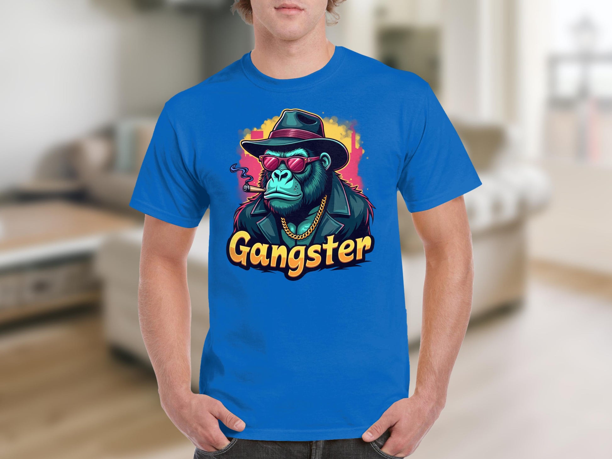 A person in a blue Garment Graphics T-Shirt, featuring urban style and a cartoon gorilla with sunglasses, hat, and gold chain. Gangster is boldly printed below. The classic fit pops against the blurry indoor background with light brown flooring.