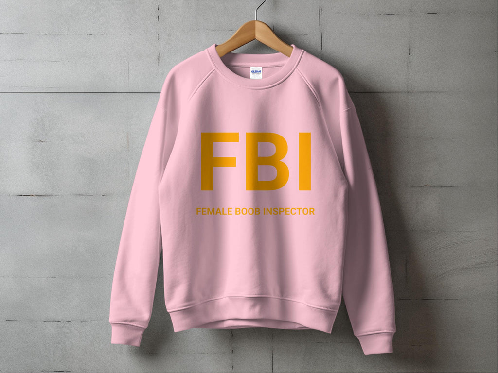 A pink Garment Graphics sweatshirt hangs on a wooden hanger against a grey wall, featuring FBI in large yellow letters with Female Boob Inspector underneath. It offers unisex sizing for all styles.
