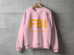 A pink Garment Graphics sweatshirt hangs on a wooden hanger against a grey wall, featuring FBI in large yellow letters with Female Boob Inspector underneath. It offers unisex sizing for all styles.
