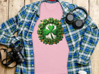 A Garment Graphics pink graphic T-shirt with a shamrock clover design sits on a wooden surface. Surrounding it are a blue plaid shirt, black headphones, a camera, and a white coffee cup lid—ideal for embracing the St. Patricks Day vibe in style.