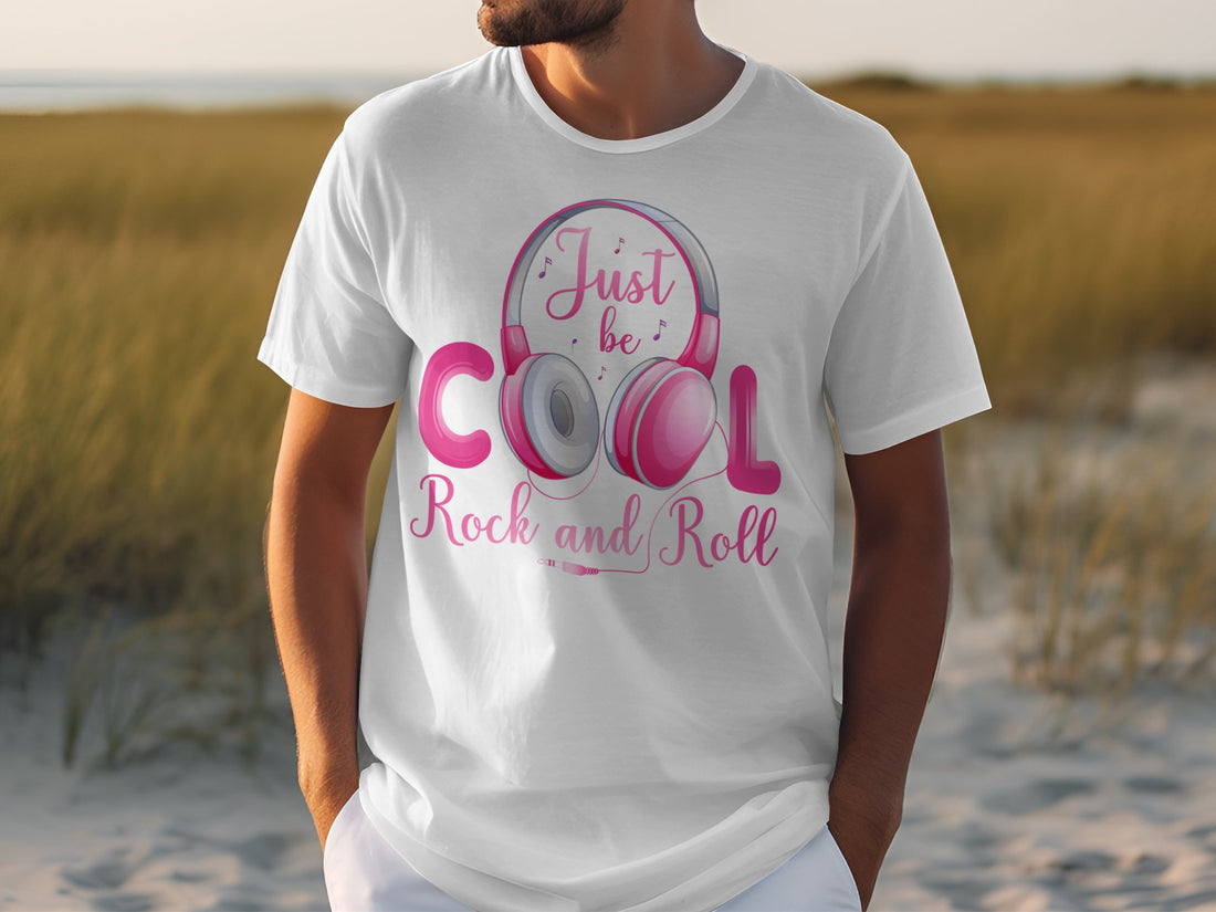 A music enthusiast wears a Garment Graphics white T-shirt with pink Fun Headphones and the text Just be Cool, Rock and Roll, standing outdoors in sand with tall grass swaying behind them.