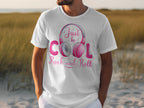 A music enthusiast wears a Garment Graphics white T-shirt with pink Fun Headphones and the text Just be Cool, Rock and Roll, standing outdoors in sand with tall grass swaying behind them.