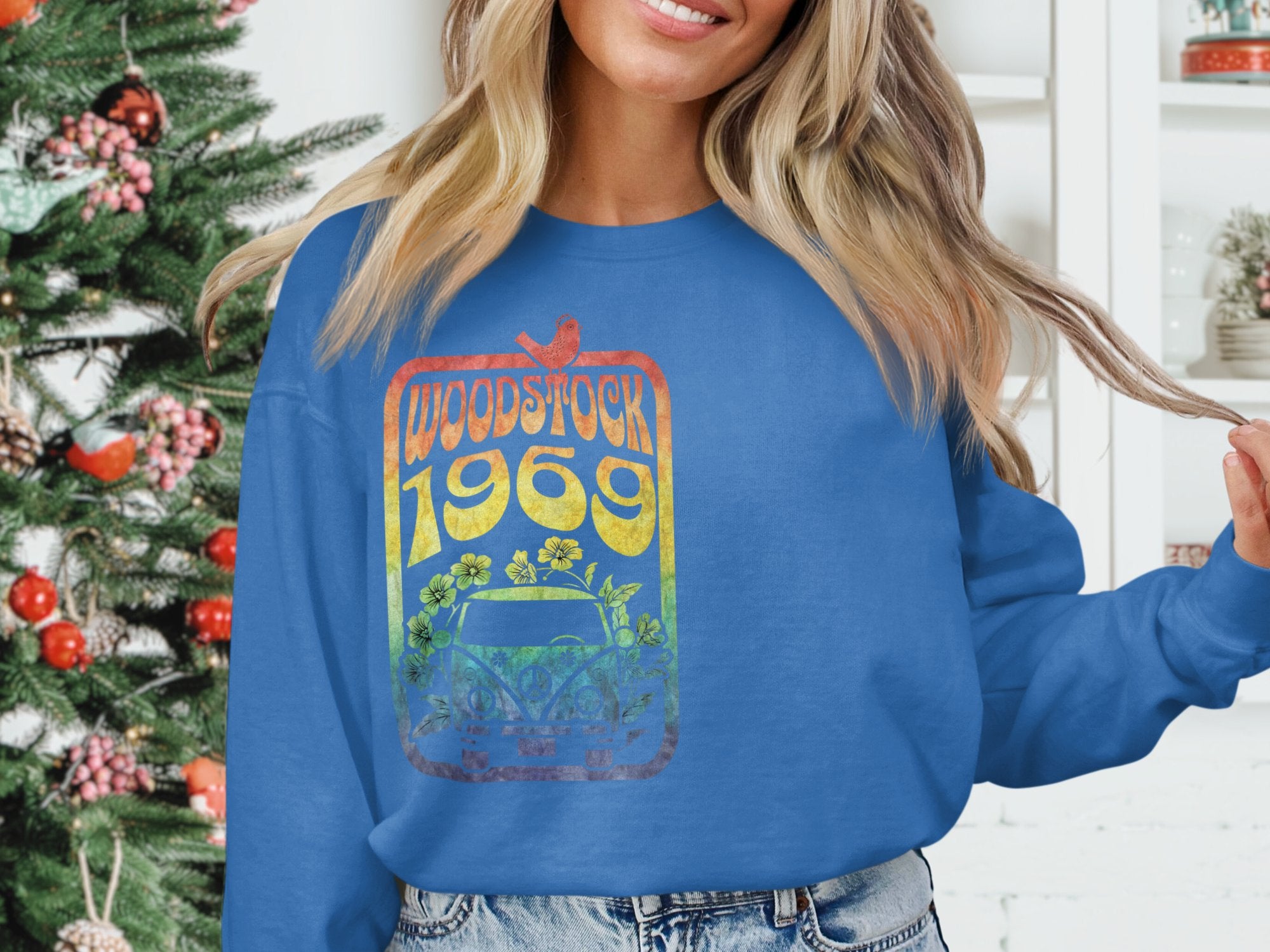 Someone in a blue Garment Graphics sweatshirt with a vintage 1969 Woodstock design stands next to a decorated Christmas tree. The retro rainbow palette, florals, and car perfectly capture the festivals spirit. The person has long hair and is partially cropped out of the frame.