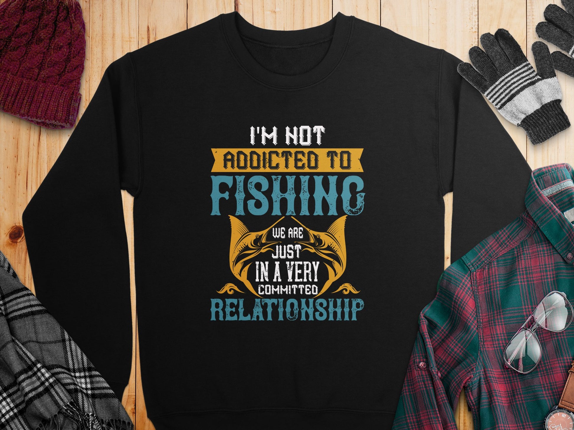 A Garment Graphics sweatshirt, lying on wood, humorously declares: Im not addicted to fishing, we are just in a very committed relationship. Its adorned with a red knit hat, black gloves, gray scarf, and plaid shirt. Unisex sizing suits anyone passionate about angling adventures.