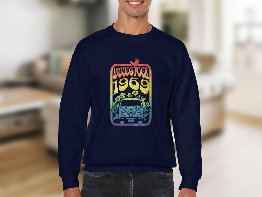 A person wearing a dark Garment Graphics sweatshirt exudes retro vibes with its vintage 1969 design, displaying the text Woodstock 1969 over a flower-adorned car and peace sign. A softly focused indoor living room in the background enhances the nostalgic atmosphere.