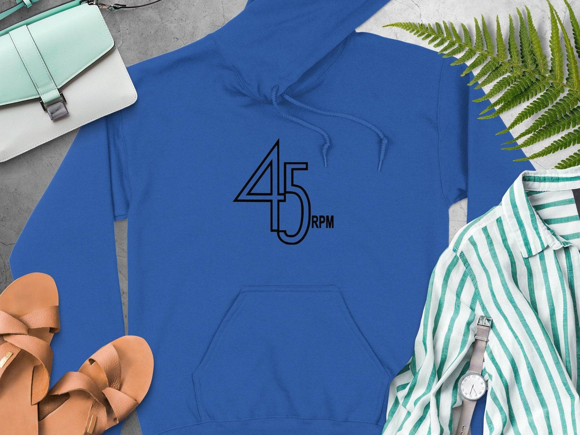 A Garment Graphics blue retro music hoodie with 45 RPM on the front is displayed on concrete, accompanied by tan sandals, a striped shirt with a watch, a green purse, and a fern leaf—ideal for vinyl record aficionados.