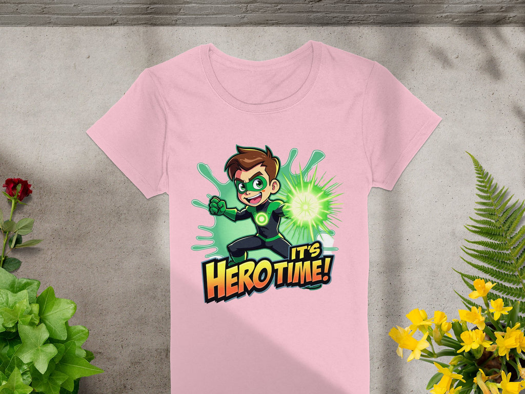 The Garment Graphics pink t-shirt showcases a young hero with a green superhero design and glowing fist, set against the phrase Its Hero Time! in bold. Lush plants surround this vibrant tee, laid out on a gray surface, making it perfect for adding color to any wardrobe.