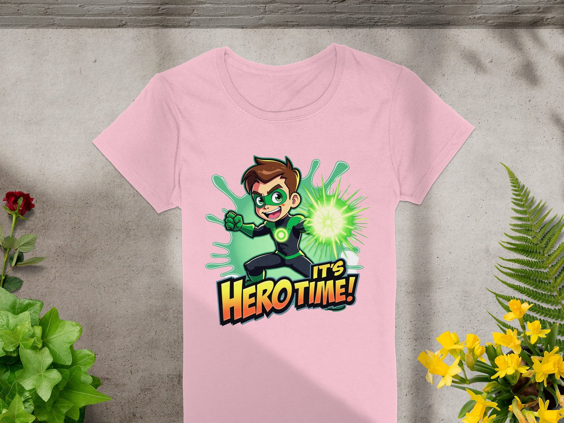 The Garment Graphics pink t-shirt showcases a young hero with a green superhero design and glowing fist, set against the phrase Its Hero Time! in bold. Lush plants surround this vibrant tee, laid out on a gray surface, making it perfect for adding color to any wardrobe.