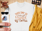 A white Garment Graphics graphic sweatshirt featuring This Day is Powered by Coffee in brown and orange lettering lies on a wooden surface, surrounded by a yellow jacket, pink loafers, sunglasses, makeup items, and a small blue pouch.