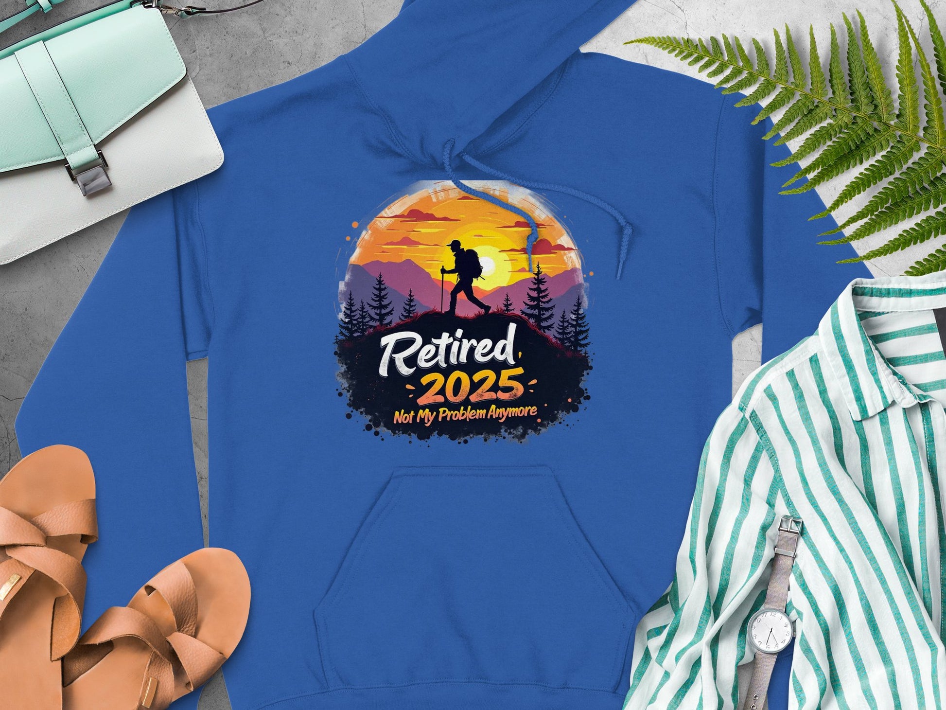 Garment Graphics blue hoodie features a sunset mountain design with a hiker and trees. It has the text Retired 2025, Not My Problem Anymore. A striped shirt, white watch, fern leaves, sandals, and bag accompany it on a grey surface.