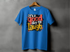 Displayed on a wooden hanger against a gray wall, Garment Graphics Its a Good Day to Laugh t-shirt bursts with vibrant colors. The cheerful message pops in red, white, black, and orange lettering, infusing joy into your day.