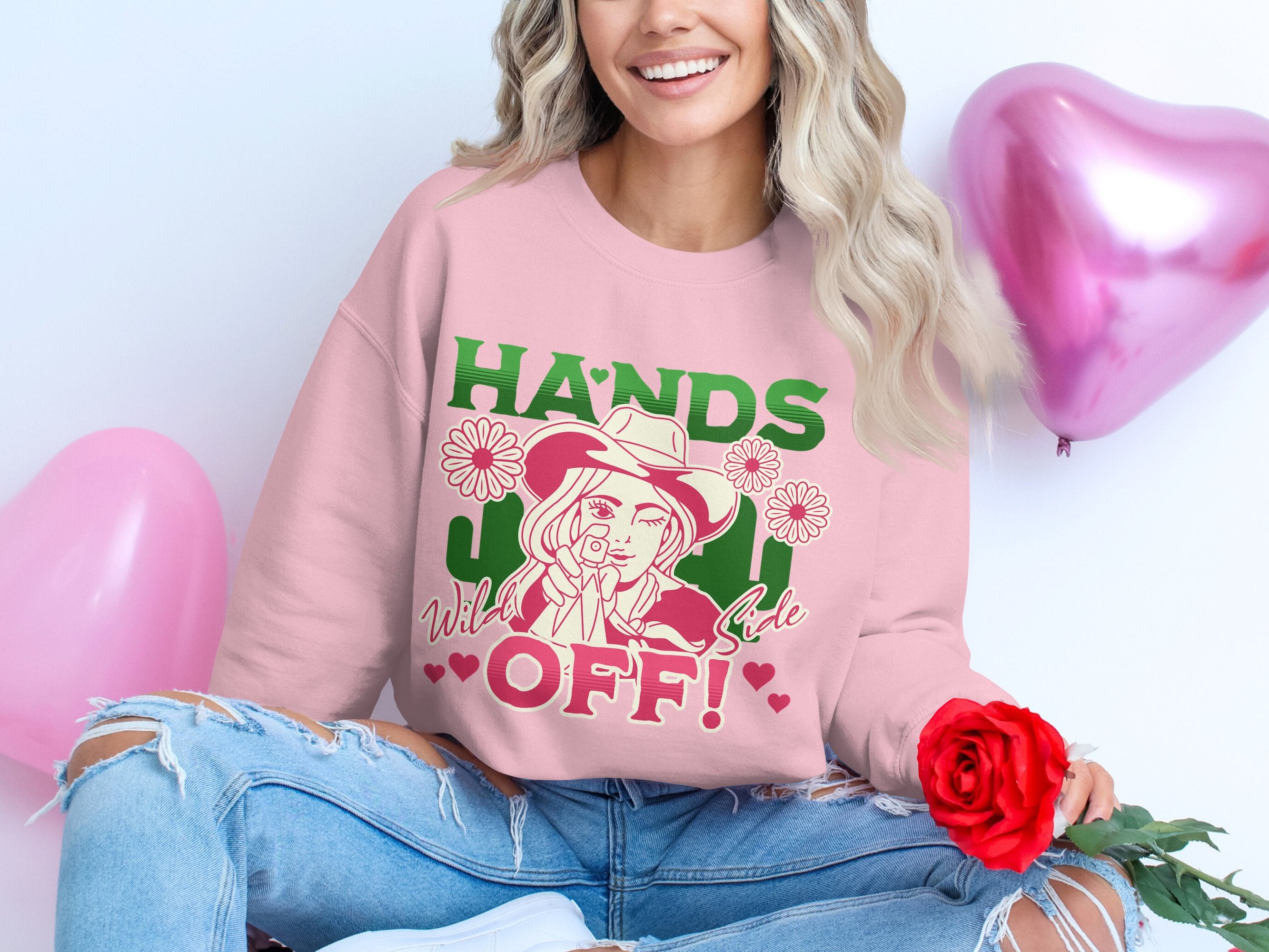 A woman leans against a white wall adorned with pink and red heart-shaped balloons, wearing a loose-fit pink Hands Off! sweatshirt from Garment Graphics Hands Off Wild Side Sweatshirts collection. She holds a red rose, perfectly complementing her ripped jeans for a chic appearance.