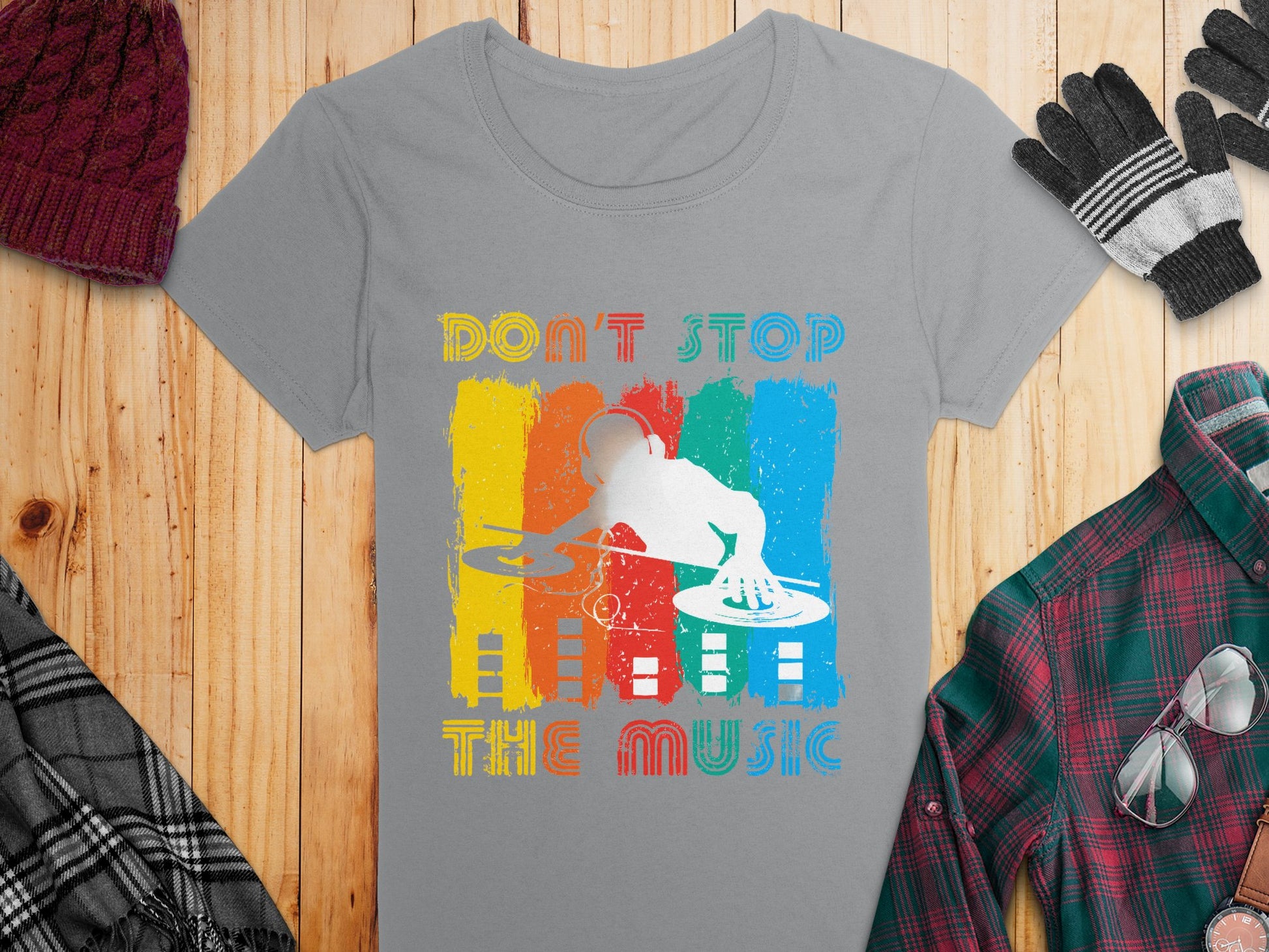 Gray Garment Graphics DJ graphic tee with a white silhouette and Dont Stop the Music text on a vibrant, distressed background. Perfect for music lovers, its displayed on wood, surrounded by a plaid shirt, gloves, scarf, and knit hat for a cozy look.