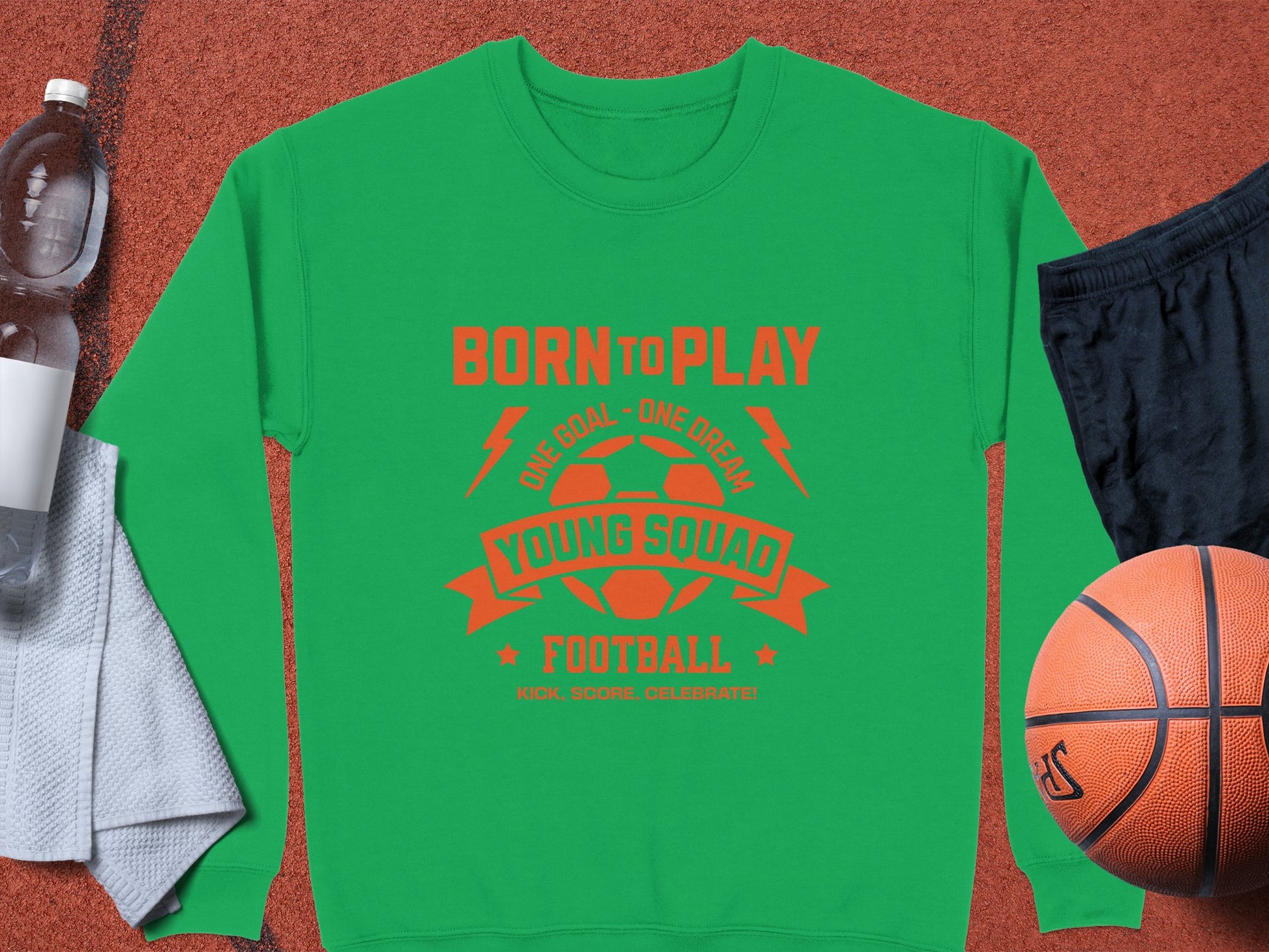 A green unisex sweatshirt by Garment Graphics, featuring an orange BORN TO PLAY and YOUNG SQUAD FOOTBALL design, is on a sports court beside a basketball, water bottle, white towel, and football—ideal for young aspiring footballers.