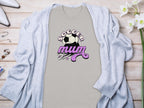 A beige classic fit T-shirt by Garment Graphics with a purple Soccer Mum design and soccer ball motif lies on a light wooden surface. Its partially covered with a light blue cardigan, accompanied by elegant white flowers, jewelry, and white high-heeled shoes.