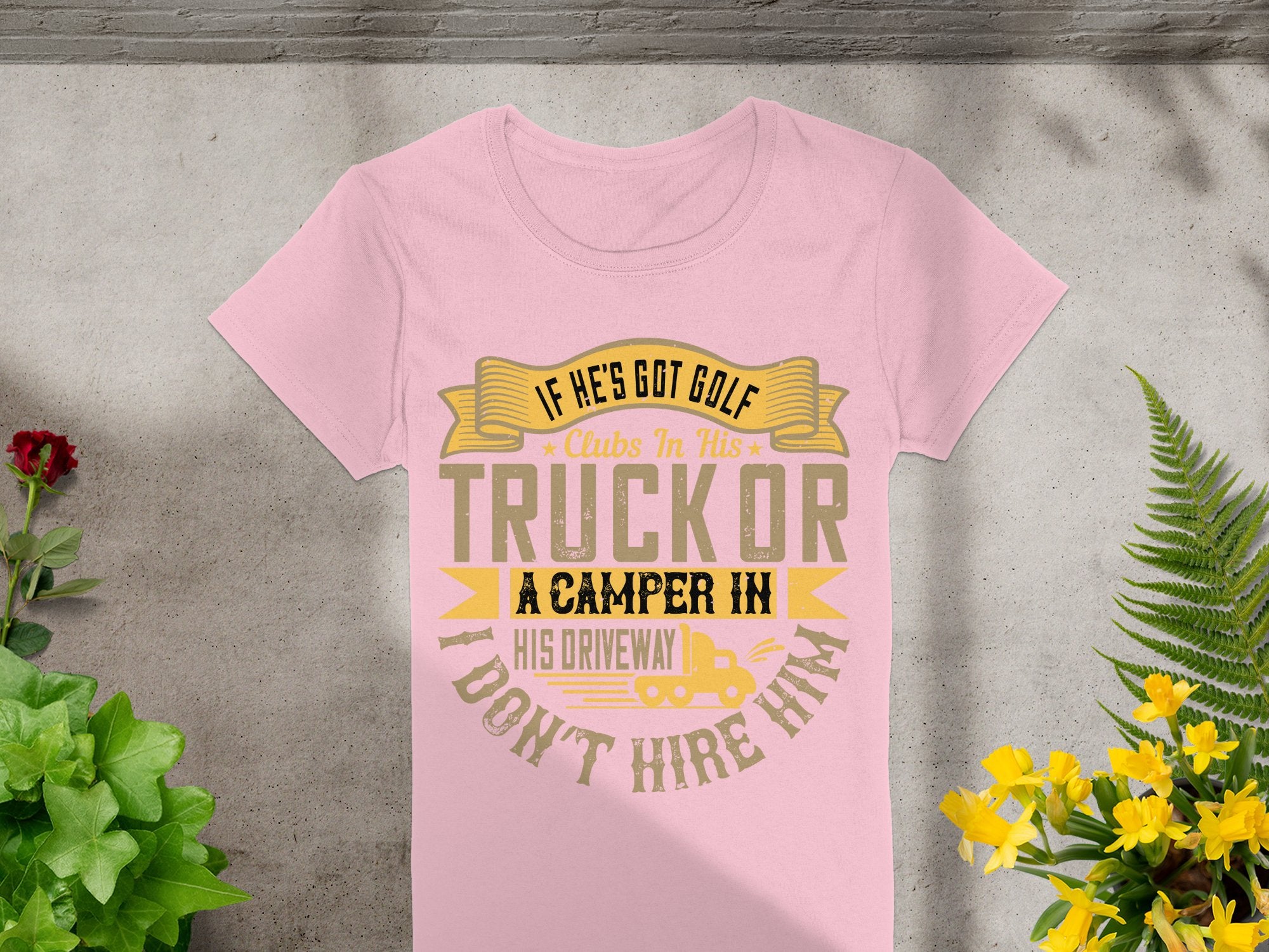 A Garment Graphics pink T-shirt, displaying a funny golf graphic and the quote If hes got golf clubs in his truck or a camper in his driveway, dont hire him, is styled on concrete with greenery and yellow flowers. Perfect for women who love quirky designs.