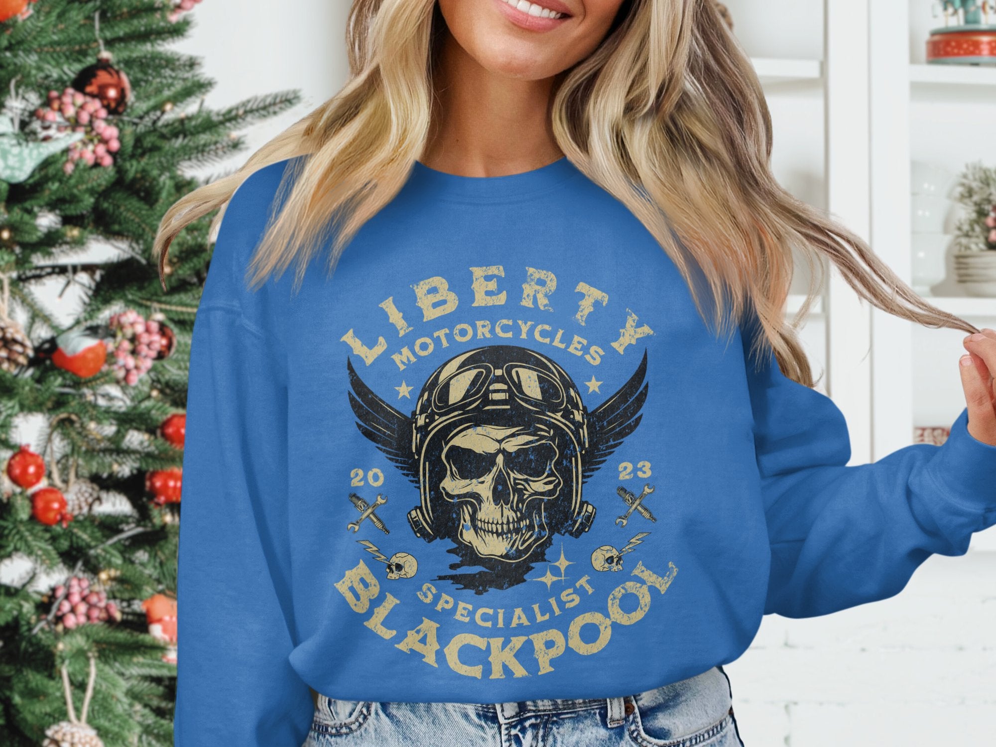 A person wears a Garment Graphics blue sweatshirt featuring a Liberty Motorcycles Specialist Blackpool 2023 design with a skull, goggles, and wings for a biker vibe. A decorated Christmas tree adds festivity in the background.