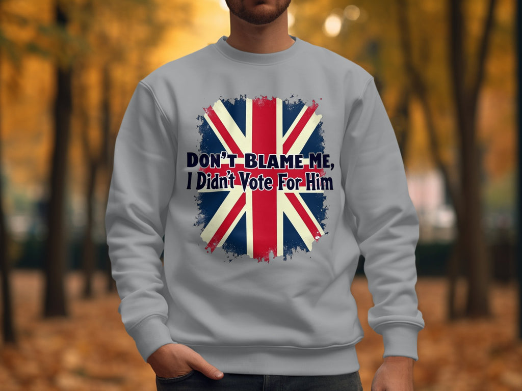 A person dons a Garment Graphics light gray sweatshirt featuring the British flag and the phrase, Dont blame me, I didnt vote for him, set against a tree-lined path with autumn foliage.
