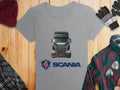 Garment Graphics gray Scania T-shirt showcases a bold black truck graphic, designed for automotive fans. Styled on wood with a red knit hat, gloves, green plaid shirt, and black-and-white checked scarf—perfect for graphic T-shirt aficionados seeking style.