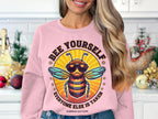 A person in a pink Garment Graphics Summer Edition sweatshirt with a cartoon bee in sunglasses and Bee Yourself, Everyone Else Is Taken! is in a festively decorated kitchen. The unisex sizing makes it perfect for everyone.