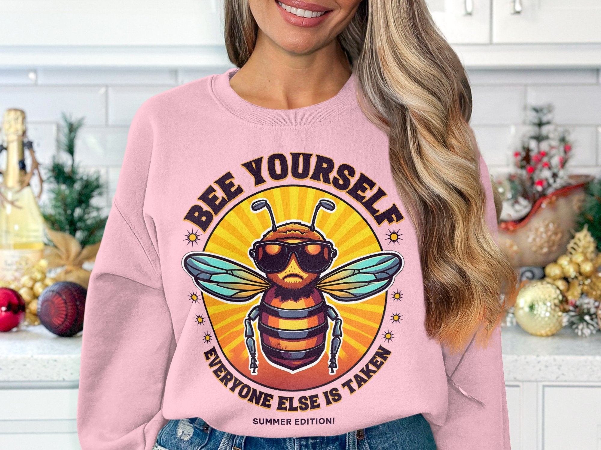 A person in a pink Garment Graphics Summer Edition sweatshirt with a cartoon bee in sunglasses and Bee Yourself, Everyone Else Is Taken! is in a festively decorated kitchen. The unisex sizing makes it perfect for everyone.