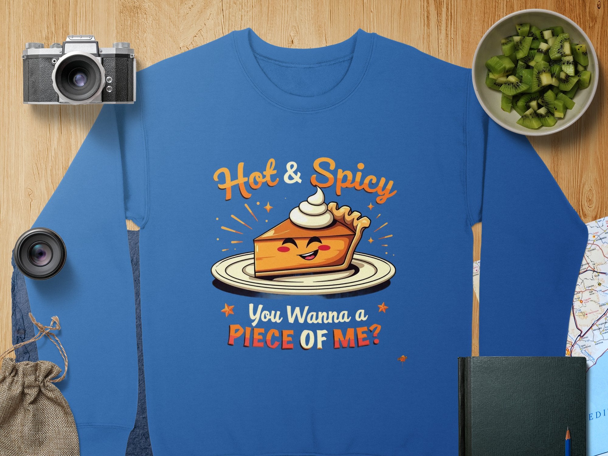 A blue Garment Graphics sweatshirt on wood shows a cartoon pie slice with whipped cream, eyes, and mouth, saying Hot & Spicy You Wanna a Piece of Me? Nearby are a camera, lens, notebook, cucumber slices, and map. This unisex piece offers playful comfort.