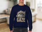 A person wearing a navy blue Garment Graphics sweatshirt with an ornate eye design above GAZE is indoors. The blurred background with neutral tones highlights the unisex appeal of this stylish garment.