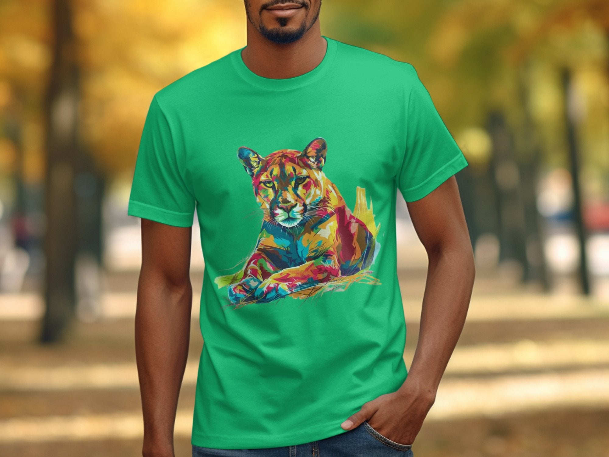 A person stands outdoors wearing a green Garment Graphics T-shirt with an abstract lion print. The vibrant artwork pops against trees with yellow leaves, evoking autumn. Their hand rests casually in their pocket.