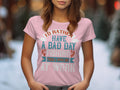 A woman stands outdoors wearing a light pink Garment Graphics fishing T-shirt for women, featuring the phrase Id rather have a bad day fishing than a good day at work in vibrant colors. The blurred background suggests an outdoor setting perfect for fishing enthusiasts.