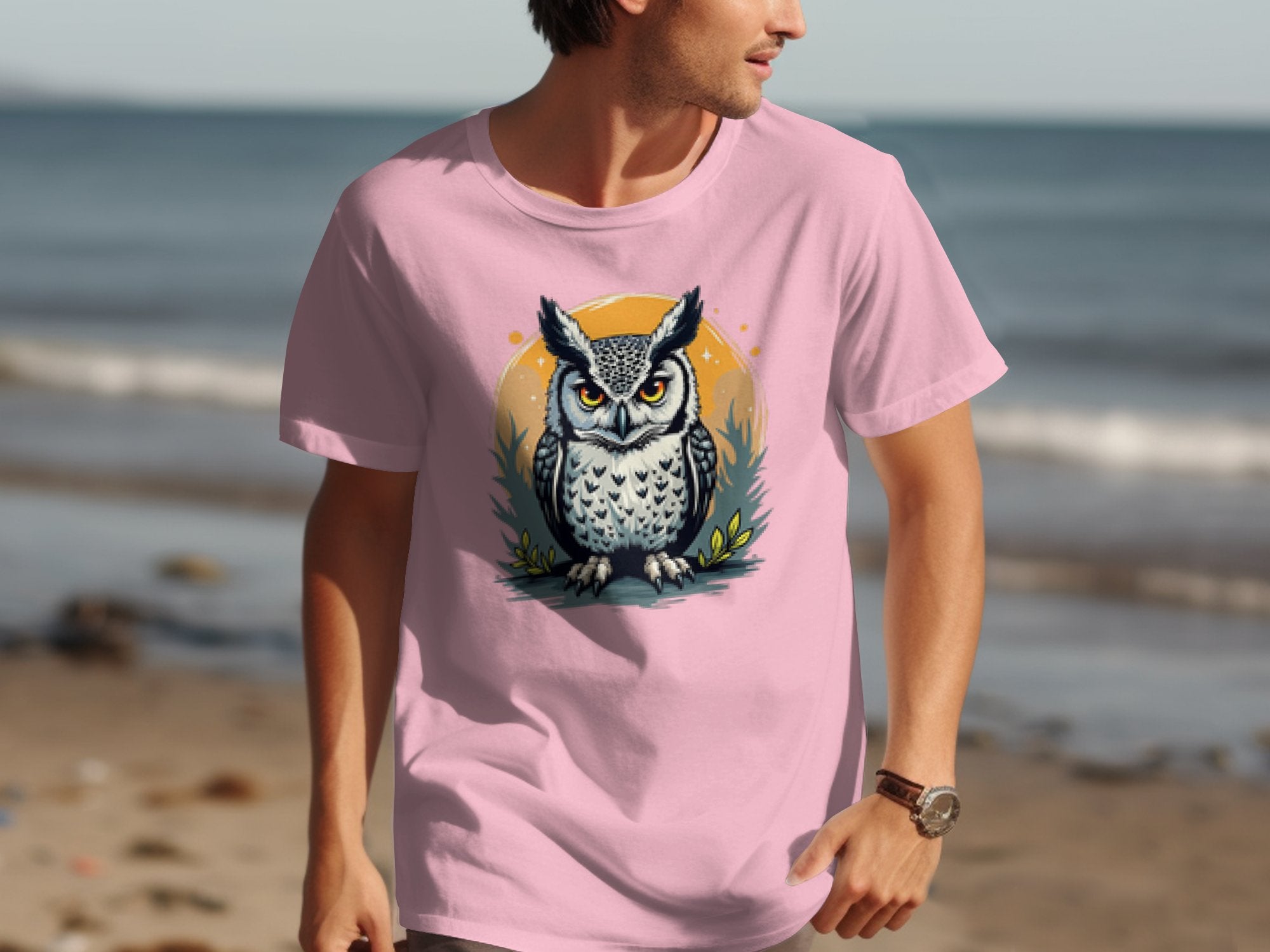 On a beach walk, someone wears a pink Garment Graphics T-shirt with an owl graphic inside a yellow circle with foliage. The sandy shore and ocean form the serene background.