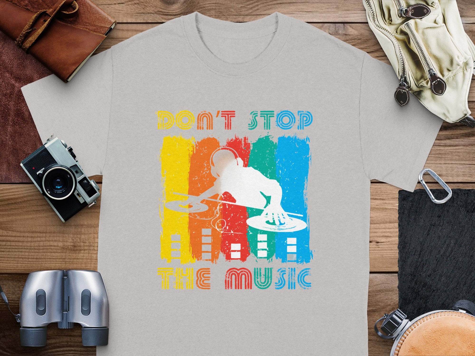 A gray t-shirt by Garment Graphics features a DJ graphic with a headphone-wearing silhouette and turntables against vibrant stripes. The phrase Dont Stop the Music stands out, making it ideal for music fans. Displayed on wood among assorted items, its perfect for lively gatherings.