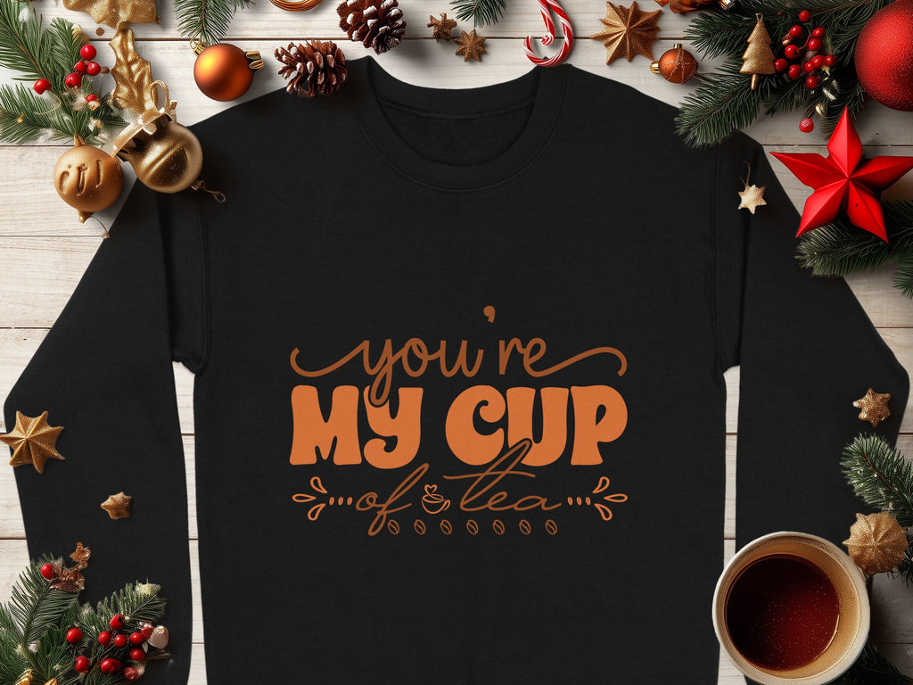 A cozy black Garment Graphics sweatshirt featuring Youre My Cup of Tea in vibrant orange text rests on a wooden surface, surrounded by festive Christmas decorations like pine cones, ornaments, stars, a candy cane, and a steaming tea cup—ideal for unisex sizing.
