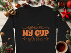 A cozy black Garment Graphics sweatshirt featuring Youre My Cup of Tea in vibrant orange text rests on a wooden surface, surrounded by festive Christmas decorations like pine cones, ornaments, stars, a candy cane, and a steaming tea cup—ideal for unisex sizing.