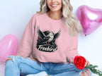 Sitting cross-legged and radiating joy, a person wears a Garment Graphics Freebird Graphic Sweatshirt featuring an eagle design. Theyre holding a red rose while two pink heart-shaped balloons float in the background.