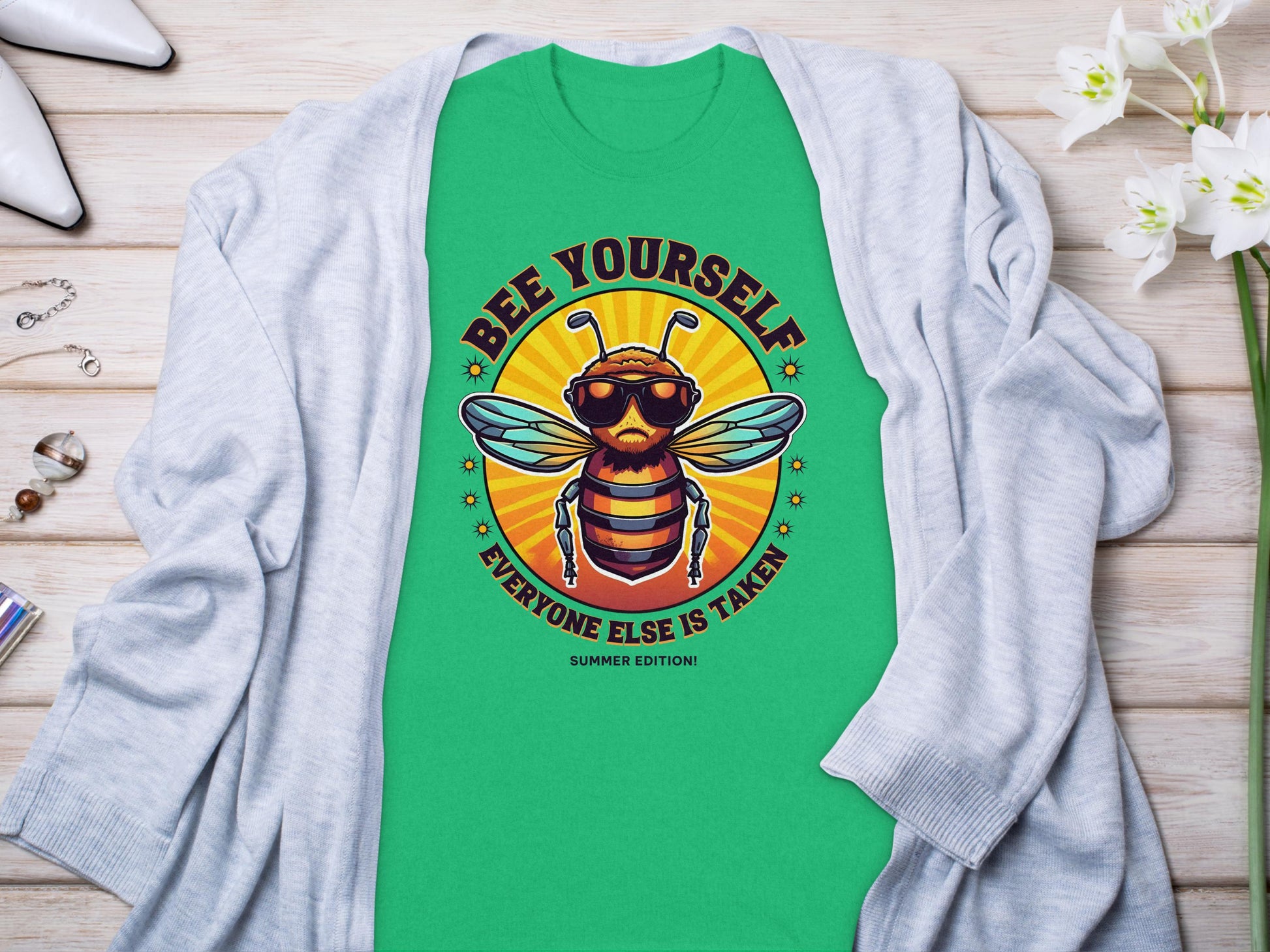 The Garment Graphics bee T-shirt, perfect for family sizing, showcases a sunburst and sunglasses-wearing bee with Every One Else Is Taken, Summer Edition text, styled with a gray cardigan and flowers on a wooden surface.