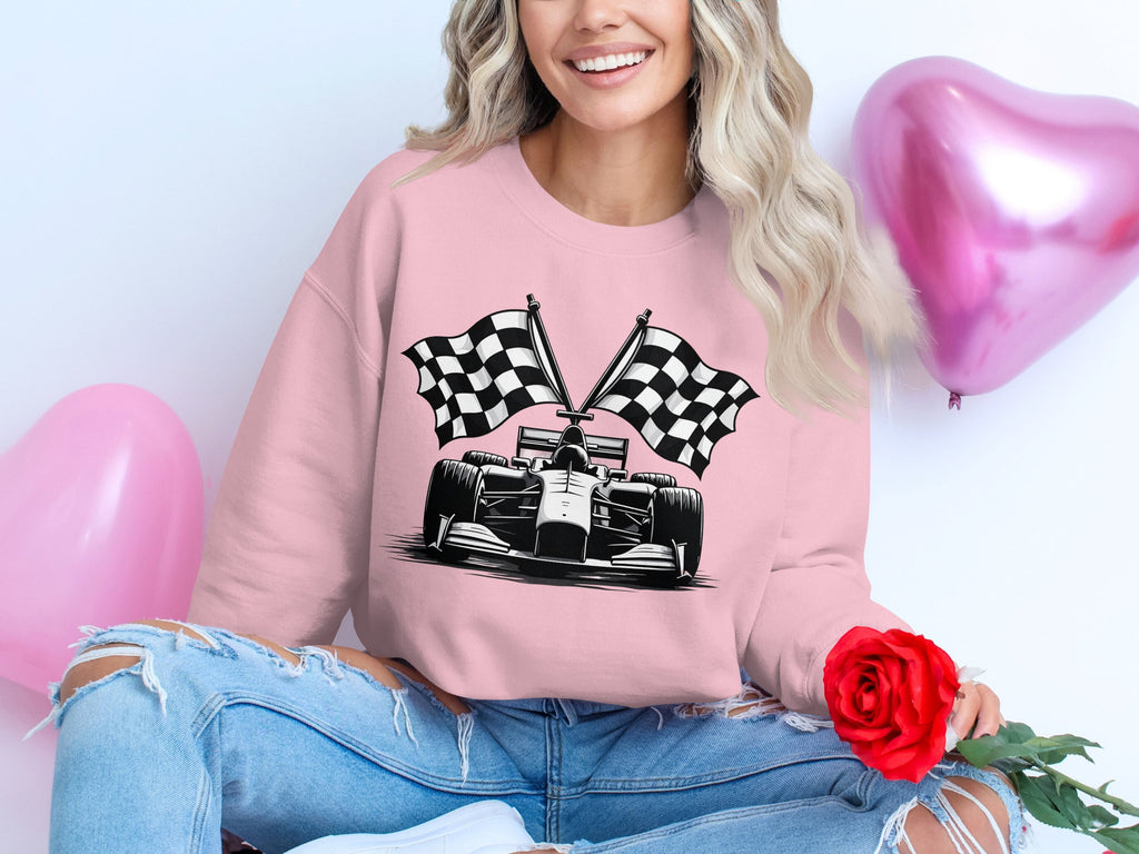 A woman sits cross-legged in a Garment Graphics pink racing car sweatshirt with checkered flag print, holding a red rose amid pink heart-shaped balloons. She smiles as her stylishly ripped jeans reveal her knees.