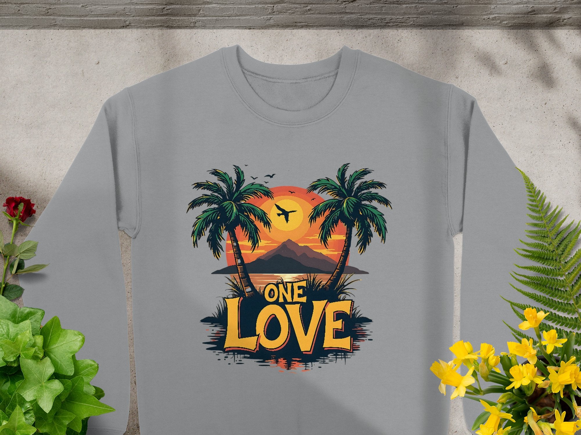 Garment Graphics gray sweatshirt showcases a One Love Sunset design with palm trees, a sunset, a plane above mountains, and is framed by green plants and yellow flowers, capturing carefree vibes.