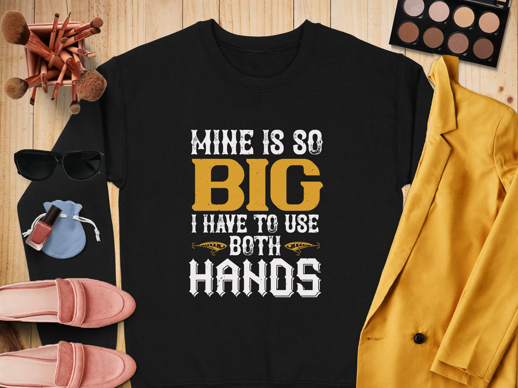 The Garment Graphics black shirt boldy states Mine Is So Big I Have To Use Both Hands in yellow and white. Its showcased on a wooden surface with makeup brushes, sunglasses, a yellow jacket, pink shoes, and makeup items.